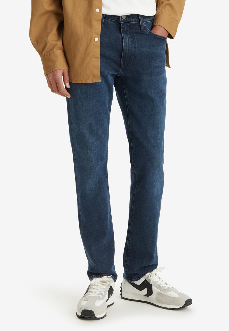 Jean en denim bleu foncé avec une coupe droite, présentant un design classique à cinq poches et des détails de couture subtils, associés à des baskets blanches.