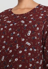 Blusa floreale bordeaux con scollo a barca. Presenta piccoli fiori bianchi e blu con accenti verdi, realizzata in tessuto leggero.
