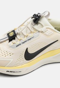 Nike Pegasus tenisice s kremastim mrežastim gornjištem, crnim swoosh-om, žutim detaljima i aerodinamičnim dizajnom s vidljivom Air Zoom jedinicom.