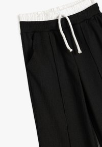 Pantalones negros con una superficie texturizada, cinturilla elástica blanca y cordón ajustable. Se pueden ver los bolsillos laterales.