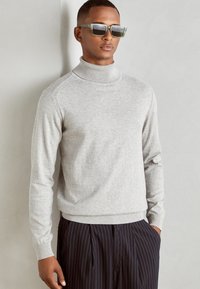 Pulover turtleneck gri deschis dintr-un material moale, tricotat, cu manșete și tiv cu nervuri, asortat cu pantaloni în dungi întunecate și ochelari de soare.