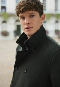 Tmavozelený vlnený kabát s vysokým golierom, dvojradovým zapínaním a čiernymi detailmi na gombíkoch, zobrazený na rozostrenom pozadí.