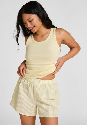 Femme souriante ajustant son haut sans manches jaune pâle, portant un short vichy jaune clair sur un fond gris uni.