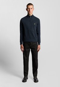Pullover blu navy con zip e lavorazione a maglia, abbinato a pantaloni in denim scuro e stivali neri con lacci. Accento del logo dorato sul maglione.