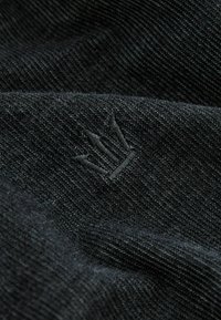Tejido negro con una textura tejida; presenta un sutil logo bordado en un tono más oscuro. Superficie suave con un ligero brillo.