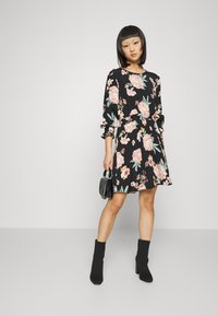 Vestido preto com flores e rosas cor-de-rosa, mangas longas, cintura franzida e saia rodada. Combinado com botins pretos e uma bolsa pequena.