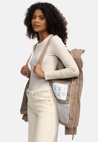 Veste zippée beige avec une poche en mesh et des détails graphiques. Le modèle porte un top côtelé beige clair et un pantalon couleur crème, présentant une tenue décontractée.
