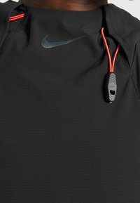 T-shirt de sport noir Nike avec logo swoosh réfléchissant près du col et cordons rouges avec un arrêt en plastique noir sur le côté droit.