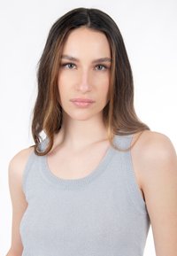 Mujer joven con cabello largo y castaño, vistiendo una blusa de punto plateada sin mangas, mirando directamente a la cámara con una expresión neutral.