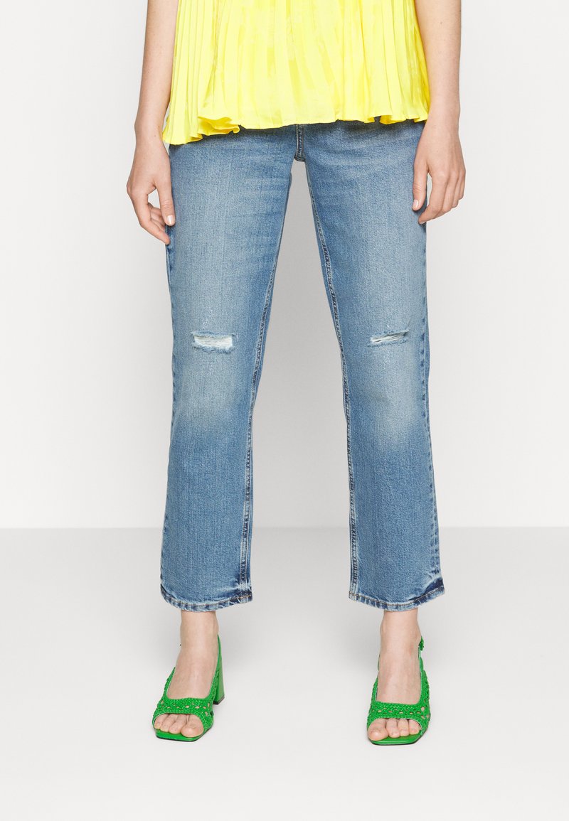 Jeans desgastados azul claros com joelhos rasgados; combinados com sandálias de salto verde com detalhes intrincados e dedos dos pés abertos. O topo é amarelo e pregueado.