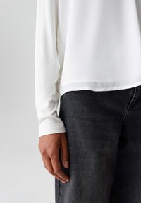 Weiße Langarmbluse mit glatter Textur, mit abgerundetem Saum, kombiniert mit dunkelgrauen Jeans, die ein lässiges Outfit betonen.