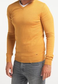 V-ringad tröja i senapsgul färg, tillverkad av mjukt stickat tyg, figursydda långärmar, ribbad fåll och manschetter, minimal logotyp längst ner.