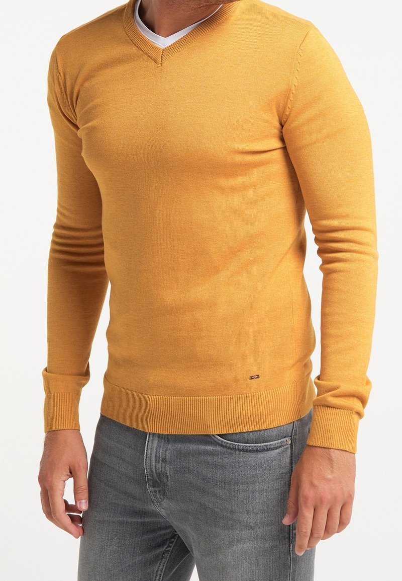 V-ringad tröja i senapsgul färg, tillverkad av mjukt stickat tyg, figursydda långärmar, ribbad fåll och manschetter, minimal logotyp längst ner.