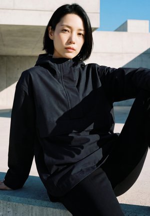 Fiatal személy rövid fekete hajjal, fekete dzsekit és nadrágot viselve, beton párkányon ül kint, modern építészet előtt.