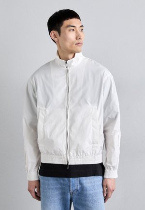 Veste bomber blanche, légère, zippée, avec un col montant, des poignets élastiques et des poches latérales, portée par-dessus une chemise noire et un jean bleu.