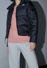 Chaqueta negra acolchada con cremallera frontal y cuello de piel sintética, usada sobre una camisa rosa claro y pantalones deportivos grises. Textura suave y acolchada.