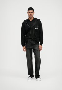 Sweatshirt com fecho éclair preto sobre uma polo preta, combinado com jeans cinzentos escuros e ténis pretos. Apresenta um bolso frontal com detalhe de logotipo.