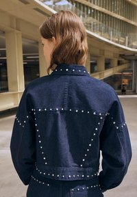 Temno modra usnjena jakna s srebrnimi žebljički ob šivih, s koničastim ovratnikom in strukturirano zasnovo, pogledana s hrbtne strani.