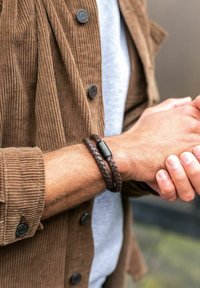 Pulsera de cuero trenzado marrón con un cierre de metal negro, llevada en una muñeca, combinada con una chaqueta de pana marrón texturizada y una camisa gris.