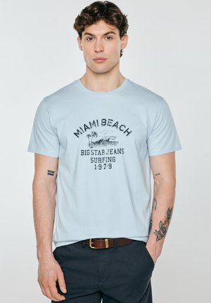 Młody mężczyzna z falowanymi włosami, ubrany w jasnoniebieski T-shirt z napisem „Miami Beach Big Star Jeans Surfing 1979” oraz ciemne spodnie, z tatuażami na lewym przedramieniu.