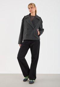 Donna che indossa un giubbotto doppiopetto grigio scuro, pantaloni neri a gamba larga e sneakers blu con lacci verde neon, in piedi contro uno sfondo bianco.