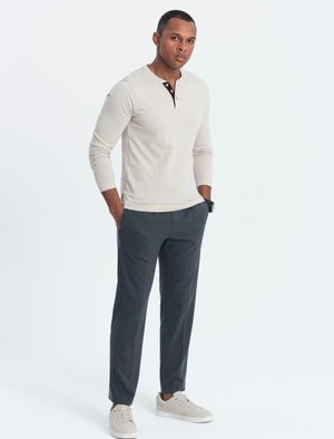Mann steht mit den Händen in den Taschen, trägt ein beiges Langarm-Henley-Shirt, dunkelgraue Hose, beiges Sneaker und eine schwarze Armbanduhr.