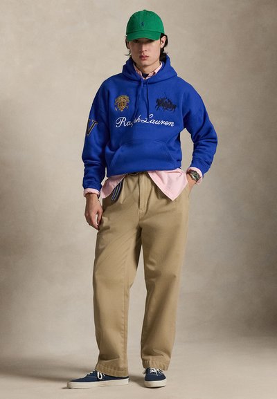 Jeune homme portant une casquette verte, un sweat à capuche Ralph Lauren bleu, une chemise rose, un pantalon beige et des baskets bleues, debout devant un fond neutre.