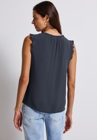 Blouse sans manches gris foncé avec des détails froncés aux épaules, texture douce et ourlet arrondi, associée à un jean clair déchiré.