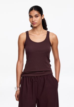 Femme aux cheveux bouclés attachés en arrière porte un débardeur marron foncé sans manches et un pantalon ample assorti, les mains dans les poches.