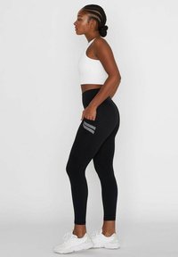 Leggings noirs avec une taille haute, une poche latérale et des accents rayés gris ; associés à un crop top ajusté blanc et des baskets blanches.