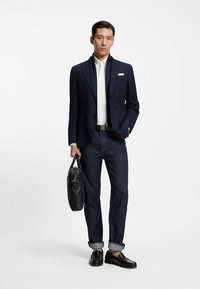 Laivastonsininen bleiseri rakenteisella kankaalla, valkoinen paita, tummat farkut, musta vyö ja mustat kengät, mustaa nahkalaukkua kantaen.