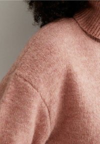 Maglione lavorato a maglia in morbido tessuto rosa, con colletto alto a coste e spalle scese. La texture appare soffice e accogliente.