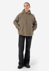 Pullover in pile color beige con zip a metà, dotato di pannelli a contrasto grigi, tasca frontale e vestibilità comoda. Abbinato a pantaloni in denim neri.