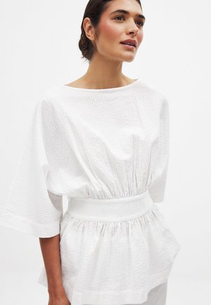 Top blanco con una tela texturizada, mangas anchas y cinturilla elastizada. Presenta una silueta suelta y drapeada, con un ligero fruncido en la cintura.