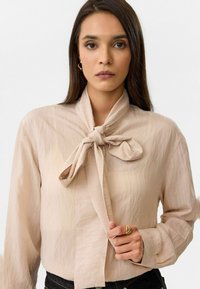 Lichtbeige blouse met lange mouwen en een strikje rond de hals, gemaakt van textuurstof. Heeft knoopmanchetten en zachte draping.