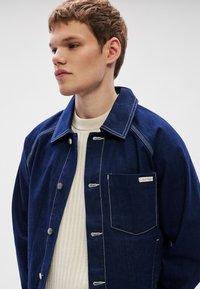 Denimjakke i mørkeblå med hvit søm, med knapper foran, brystlomme og merkeetikett. Brukt over en kremfarget ribbestrikket genser.