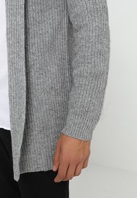 Cardigan grigio lavorato a maglia con un motivo testurizzato, maniche lunghe e apertura frontale. Il materiale sembra spesso e caldo, adatto per il clima più freddo.