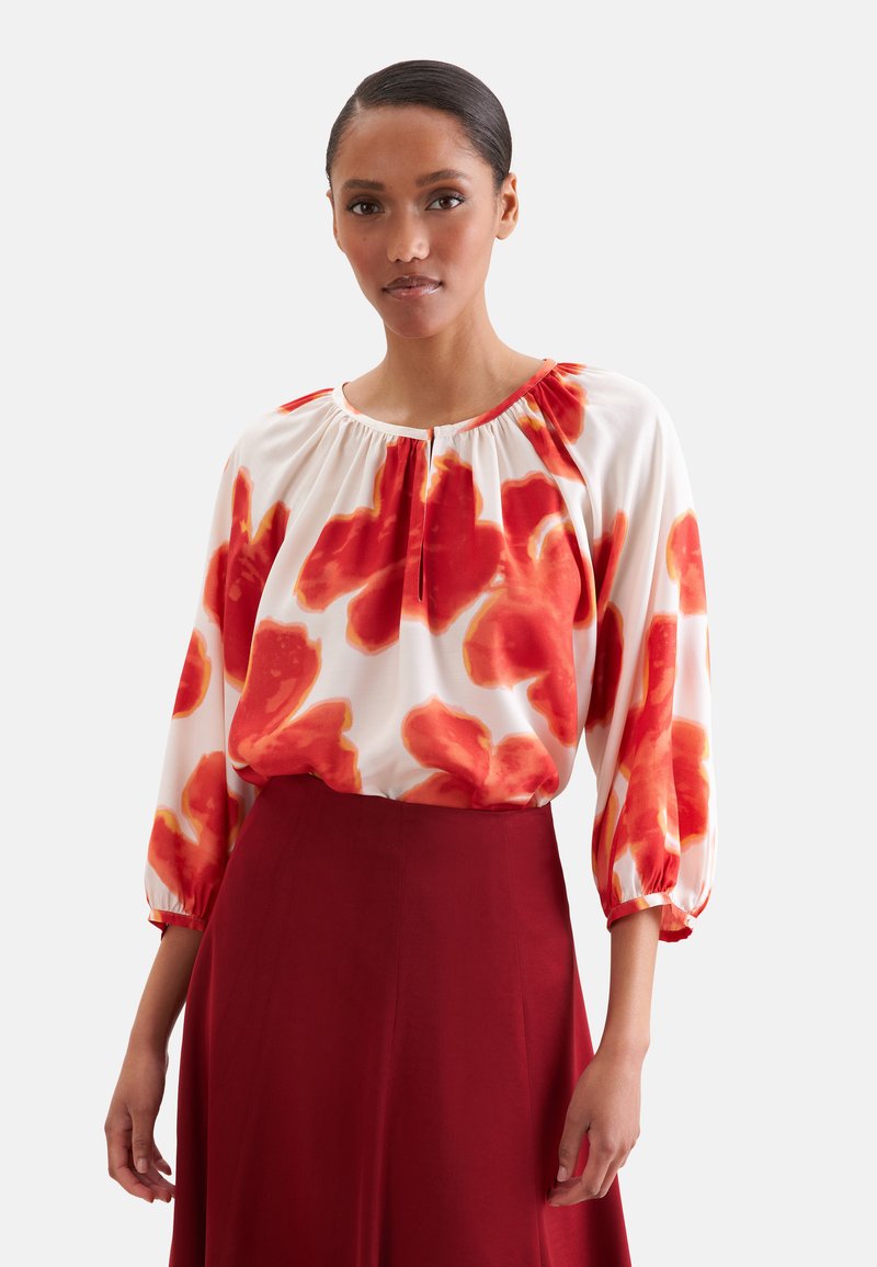 Femme portant un chemisier blanc à grands motifs floraux rouges et une jupe rouge taille haute, debout devant un fond uni.