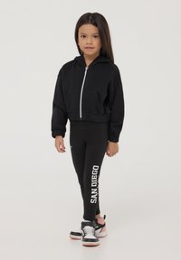 Felpa nera con zip e tasche frontali, abbinata a leggings neri con il testo 'SAN DIEGO'. Completata con scarpe da ginnastica nere e rosa.