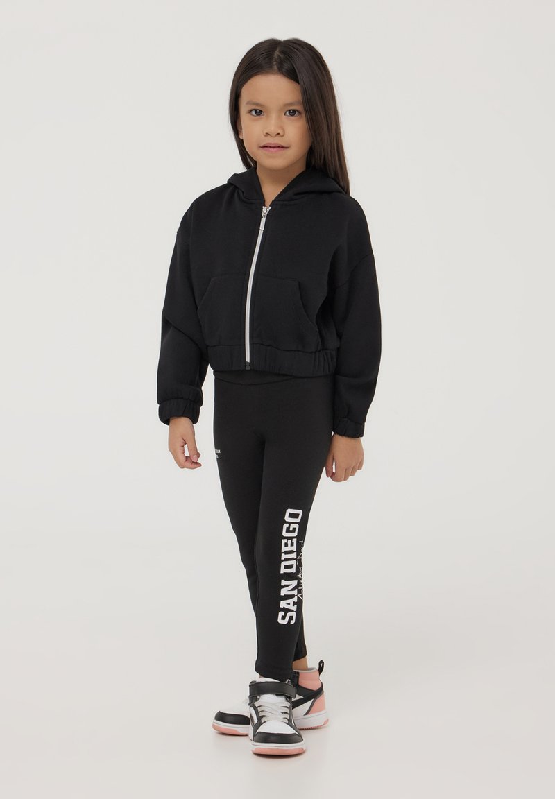 Felpa nera con zip e tasche frontali, abbinata a leggings neri con il testo 'SAN DIEGO'. Completata con scarpe da ginnastica nere e rosa.