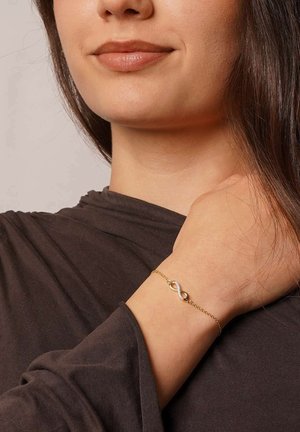 Gold chain bracelet featuring an infinity symbol with embedded gemstones, worn on a wrist against a dark fabric background.