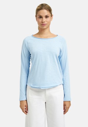 Frau steht mit Blick nach vorne, trägt ein hellblaues langärmliges Hemd und weiße Hose vor einem schlichten Hintergrund.