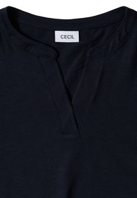Marinblå skjorta med V-halsringning och en vit etikett med texten "CECIL" sydd på insidan av kragen.