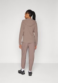 Bruine lichtgewicht hoodie met capuchon, elastische manchetten en accenten op de rug. Draag een bijpassende bruine tapered broek en zwarte sneakers.