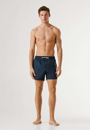 Nuori mies seisoo paljain jaloin, yllään tummansiniset uimashortsit, joissa on valkoinen kiristysnauha, valkoista taustaa vasten.