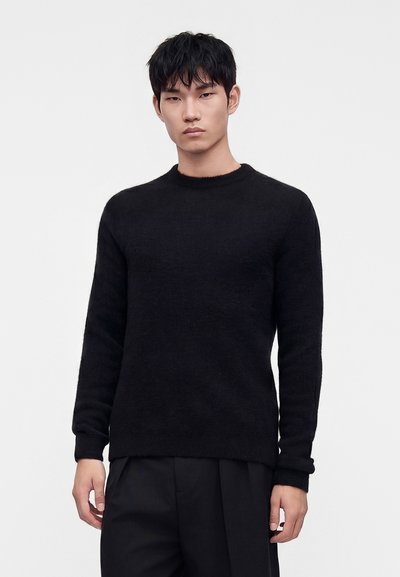 Junger Mann mit kurzen schwarzen Haaren, der einen schlichten schwarzen Langarmpullover und schwarze Hose trägt, vor einem weißen Hintergrund stehend.