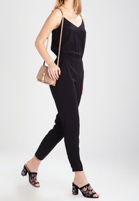 Kvinna som bär en svart ärmlös jumpsuit, en naken crossbody-väska och svarta klackmules med färgglad blommig brodyr, går.