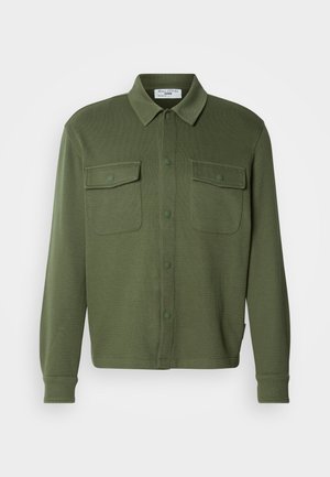 Chemise boutonnée vert olive en tissu texturé, dotée de deux poches poitrine et d'un col avec fermeture par boutons.