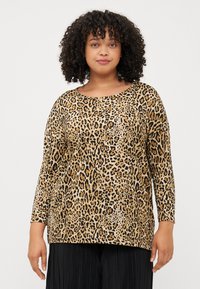 A woman with curly hair wears a long-sleeve, leopard print top. She stands against a plain background, smiling calmly.