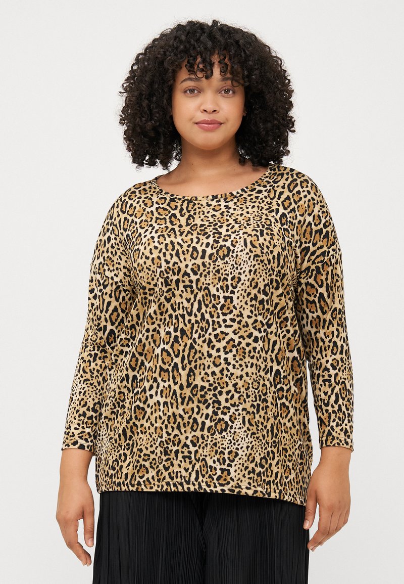 A woman with curly hair wears a long-sleeve, leopard print top. She stands against a plain background, smiling calmly.